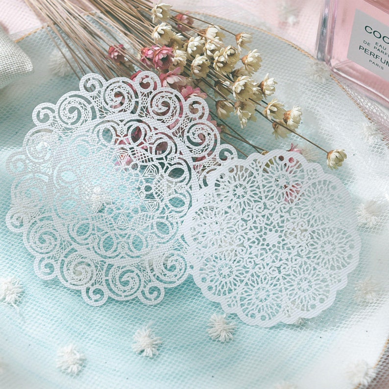 May include: Two white, intricately designed paper doilies with a delicate, lace-like pattern. The doilies are circular with scalloped edges. Dried flowers and a perfume bottle are in the background, suggesting a vintage or craft theme.