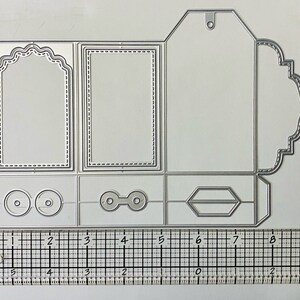 Puede incluir: Un troquel de metal plateado para manualidades, con varias formas que incluyen marcos, etiquetas y bordes decorativos. El troquel se muestra con una regla para la escala, con medidas en centímetros.