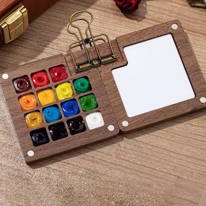 Könnte beinhalten: Ein Aquarellfarbkasten aus Holz mit Scharnierdesign. Das Set enthält 12 leuchtende, quadratische Farben, darunter Rot, Gelb, Blau und Grün. Ein weißer Mischbereich ist enthalten. Eine goldfarbene Klammer hält das Set geschlossen.