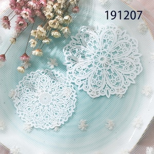May include: Two white, intricately designed paper doilies with a floral, mandala-like pattern. The doilies are on a light blue surface with a delicate, lacy texture. The number "191207" is visible in the upper right corner. Dried flowers are in the background.