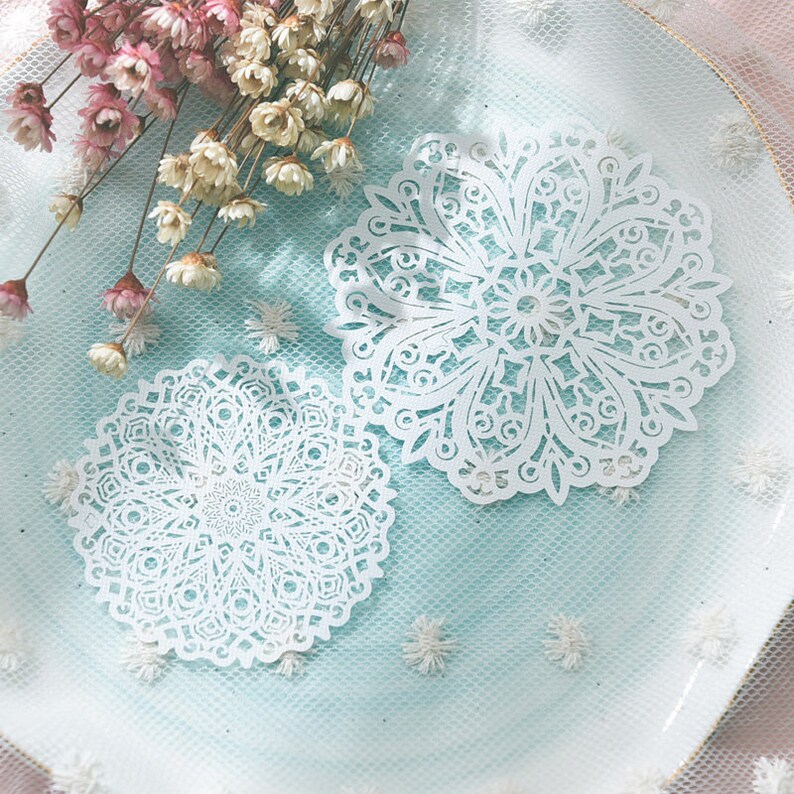 May include: Two white, intricately cut paper doilies with a floral mandala design are displayed on a light blue surface. Dried pink and white flowers are arranged in the background, adding a delicate touch to the composition. The doilies are round with detailed patterns.
