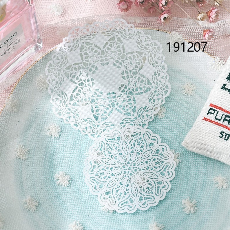 May include: Two white, intricately designed paper doilies with a delicate lace pattern. The doilies are placed on a light blue surface, with the number "191207" visible. The overall aesthetic is soft and elegant.