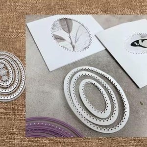 Puede incluir: Troqueles metálicos ovalados de diferentes tamaños, con bordes decorativos. Los troqueles son plateados y están colocados cerca de tarjetas blancas con recortes ovalados, uno con un diseño de hojas y otro con una mariposa. El fondo es de color neutro.