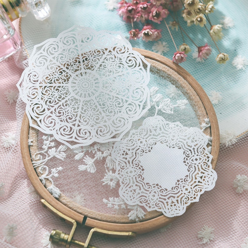 May include: Two white, intricately cut paper doilies with floral and swirl patterns rest on a wooden embroidery hoop. The doilies are placed on a sheer, embroidered fabric, with dried flowers in the background.