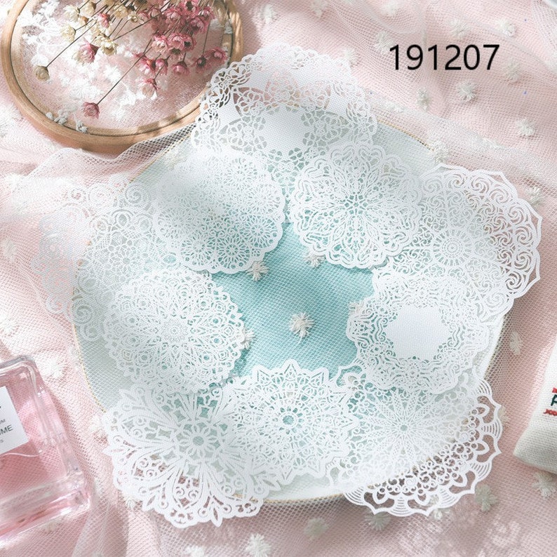 May include: A collection of white, intricately designed paper doilies arranged on a light blue plate. The doilies feature delicate, symmetrical patterns. The number "191207" is printed in the upper right corner. The image has a soft, romantic aesthetic.