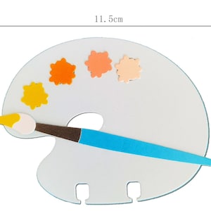 May include: A white artist's palette with a blue handle brush and colorful paint splotches. The palette measures 11.5 cm wide and 8.4 cm high. The brush has a brown handle and a yellow and white brush head. The paint splotches are yellow, orange, and peach.
