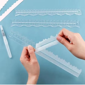 May include: Clear plastic cake decorating rulers with scalloped edges and measurement markings. A clear watercolor brush is also present. The rulers are being held by a person's hands. The background is a light blue.