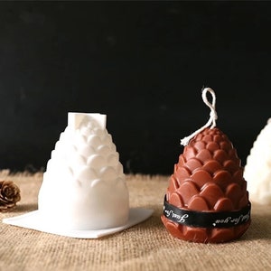 Könnte beinhalten: Zwei Kerzen in Kiefernzapfenform, eine weiße und eine braune. Die braune Kerze hat ein schwarzes Band mit dem Text "Just for you" darum gebunden.