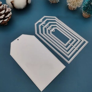 Puede incluir: Un conjunto de recortes de etiquetas de regalo de papel blanco con bordes cosidos, junto con un conjunto de troqueles de etiquetas metálicas anidadas. Los troqueles son de varios tamaños, perfectos para proyectos de manualidades y álbumes de recortes. Las etiquetas tienen forma rectangular.