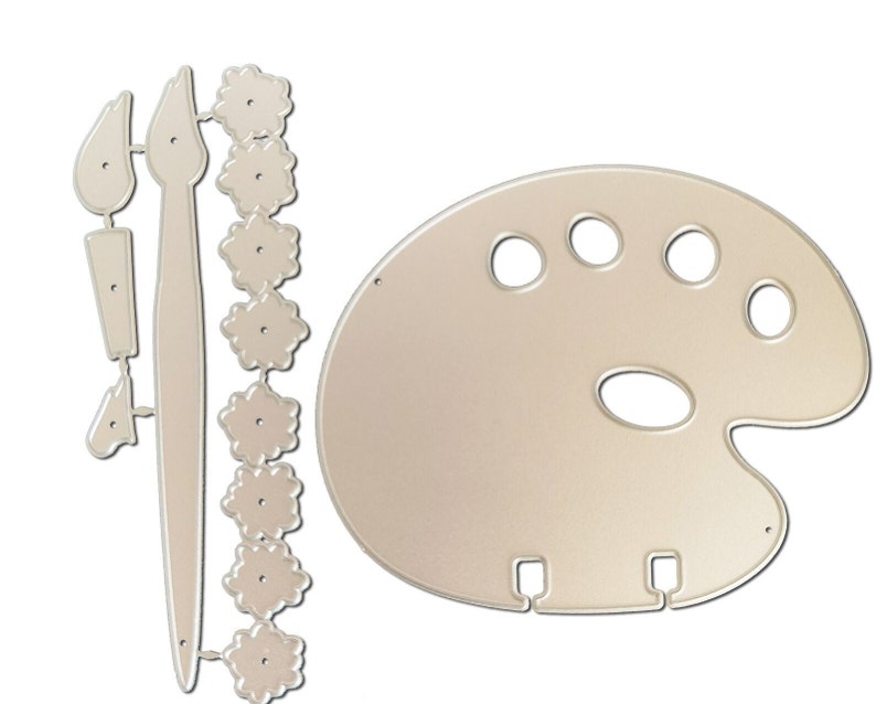 May include: A die-cut set of art-themed shapes in a light beige color. The set includes a painter's palette with oval and round paint wells, two paintbrushes, and a string of flower-shaped embellishments. The shapes are designed for crafting and art projects.
