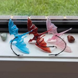Könnte beinhalten: Drei kleine Drachenfiguren mit Flügeln aus durchsichtigem Kunststoff. Die Drachen sind blau, rot und pink.