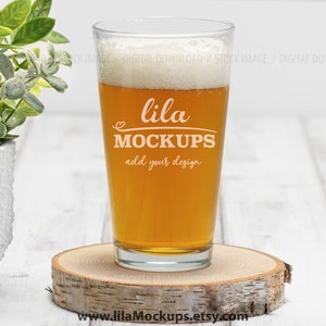 May include: A clear glass pint glass filled with a light amber beer. The glass has the text "lila MOCKUPS add your design" on the side. The glass is sitting on a wooden coaster.