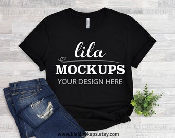 May include: Black t-shirt mockup with the text "lila MOCKUPS YOUR DESIGN HERE" on a white wooden background. The t-shirt is laying flat with a pair of blue jeans and a small green plant beside it.