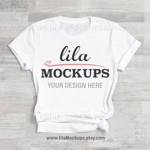 May include: White t-shirt mockup with the text "lila MOCKUPS" and "YOUR DESIGN HERE" printed on the front. The shirt is crumpled and lying on a wooden surface.