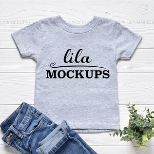 May include: A heather gray t-shirt with the text "lila MOCKUPS" in black script. The shirt is paired with blue denim jeans. A small green plant is in the bottom right corner. The shirt is a mockup for a clothing design.