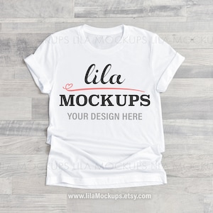 Könnte beinhalten: Weißes T-Shirt-Mockup mit dem Text "lila MOCKUPS" und "YOUR DESIGN HERE" auf der Vorderseite. Das Shirt liegt flach auf einer hellgrauen Holzoberfläche.