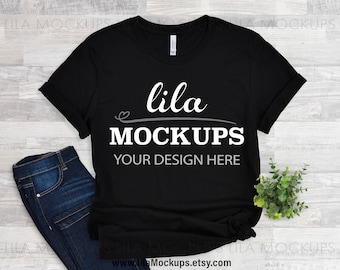 May include: Black t-shirt mockup with the text "lila MOCKUPS YOUR DESIGN HERE" on a white background. The t-shirt is laying flat on a light wood surface with a pair of blue jeans and a small green plant.