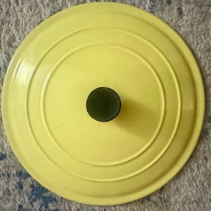 May include: A vibrant yellow pot lid featuring concentric circles and a dark green knob. The round lid, likely made of enamelled cast iron, is a kitchenware item. The lid's design is simple and functional.
