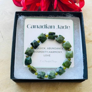 Op de afbeelding: Een groene jade armband met een vierkante vorm. De armband zit in een zwarte doos met een witte kaart waarop "Canadian Jade" en "Geluk, overvloed, voorspoed, harmonie, liefde" staat.