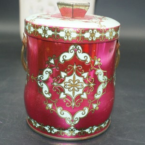 May include: A vintage red metal container with a lid and handles. The container is adorned with a white and gold ornate design. The lid has a small, gold-colored handle. The container is likely used for storage.
