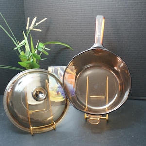May include: A vintage amber-tinted glass saucepan with a long handle and matching lid. The lid and pan are displayed on gold-colored metal stands. The cookware is set against a dark background, suggesting a kitchen or dining setting.