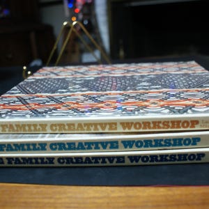 May include: Three vintage books titled "The Family Creative Workshop" are stacked. The top book has a patterned cover in shades of blue, orange, and white. The book titles are in gold and blue lettering.