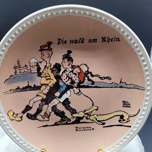 May include: A decorative plate with a pink center featuring a cartoon illustration of two people walking with a dog. The plate has a white beaded edge and the text "Die walk am Rhein". Signed by Norman Rockwell and Bill Dean Horn.