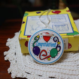 May include: A round, white Congratulations! gift tag with colorful balloons and stars, attached to a yellow box. The tag has a blue border and the word "Congratulations!" in the center. A white ribbon is attached to the tag.