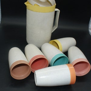May include: A vintage beige speckled plastic pitcher with a yellow lid and handle, alongside six matching tumblers. The tumblers have colored rims in shades of peach, teal, and orange. A retro kitchenware set.