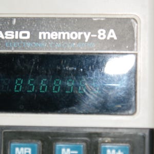 May include: Close-up of a vintage Casio memory-8A calculator. The display shows the number 856898 in green digits. The calculator has a gray and black color scheme with visible wear and tear.