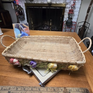 May include: Rectangular woven basket with rope handles, decorated with colorful fabric flowers. The basket is displayed on a wooden table with a vintage ruler and a framed picture. The basket measures approximately 12 inches long.