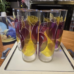 May include: Set of four vintage highball glasses with a fruit design. The glasses feature a clear body with a pattern of purple, yellow, and gold fruit and leaves. They are displayed on a cream-colored tray with silver handles.