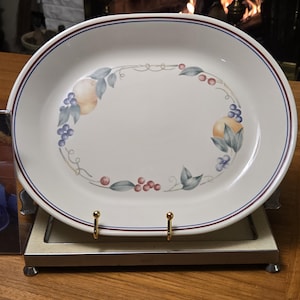 May include: Oval serving platter with a white background, featuring a fruit and vine design. The rim has a red and blue border. The platter is displayed on a metal stand with gold-colored supports.