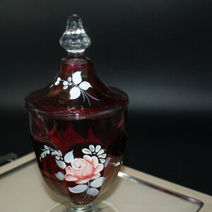 May include: A ruby red glass candy dish with a clear, faceted lid. The dish is decorated with hand-painted white flowers and a pink rose. The candy dish sits on a silver tray, against a dark background.