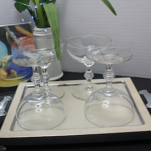 May include: A set of four clear champagne glasses on a silver serving tray. The glasses have a classic coupe shape with a decorative stem. A vintage-style print is visible in the background.