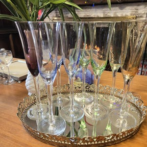 May include: A collection of champagne flutes in various colors, including purple, blue, and green, are arranged on a mirrored, ornate, gold-toned tray. The flutes are tall and slender, reflecting the light. The tray sits on a wooden surface.