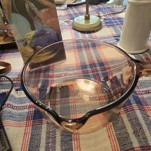 May include: A clear, light brown tinted glass saucepan with a rounded bowl, a small pouring spout on the rim, and a single handle extending from the side. The glass is transparent, showing the interior base and the smooth, curved edges of the vessel.