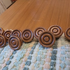 May include: A collection of eleven wooden cabinet knobs. Each knob is circular with concentric rings and a central indentation. The knobs are a warm brown color and appear to be vintage or antique. They are arranged on a multi-colored woven mat.