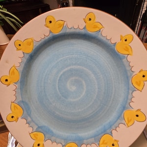 May include: A white ceramic plate with a light blue center and a border of yellow ducklings swimming in a wavy pattern. The plate is supported by a gold-colored stand.