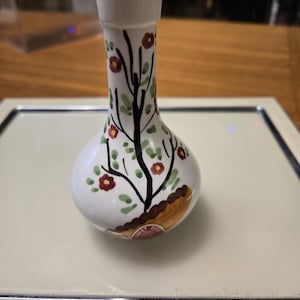 May include: A small, white ceramic vase with a bulbous body and a narrow neck. The vase is decorated with a black tree design, red flowers, and green leaves. A brown and tan patch is at the base of the tree. A small label is visible.