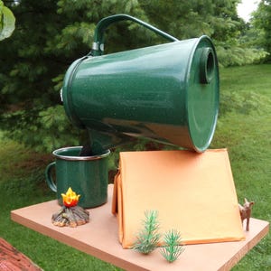 May include: A green speckled camping-style coffee pot pouring into a matching mug. A small campfire figurine, a miniature tent, and a small deer figurine are on a wooden surface. Two small green plastic pine trees are also on the surface.