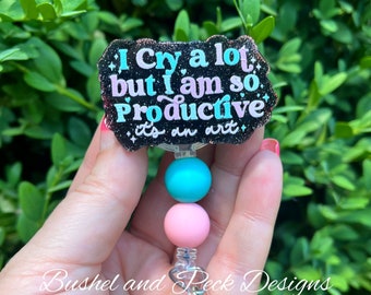 May include: A black glitter badge holder with the text "I Cry a lot but I am so Productive it's an art" in pink and blue lettering. The badge holder has a clear plastic retractable badge clip with a turquoise blue, pink, and white beaded chain.