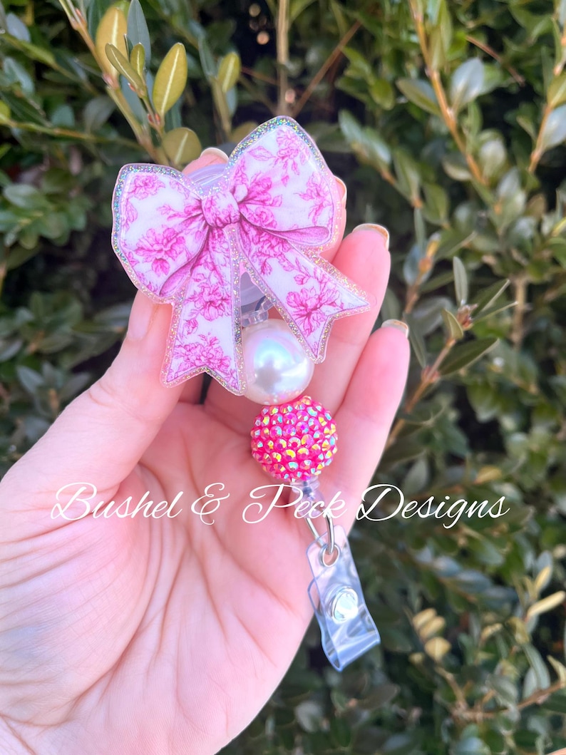 Pu&ograve; includere: Fiocco floreale rosa e bianco con una perla e un abbellimento in strass rosa attaccato a un porta badge in plastica trasparente. Bushel & Peck Designs.