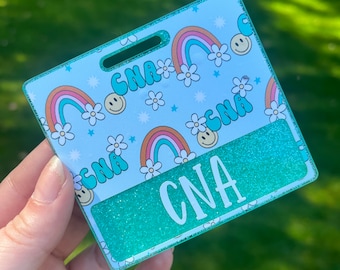 May include: A teal blue and white glitter badge with a rainbow and flower pattern. The badge has a clear plastic window with the word "CNA" printed in white.