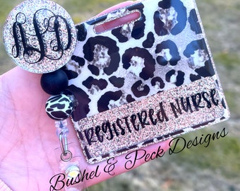 May include: A leopard print badge holder with a pink glitter background and the text "Registered Nurse" in black. The badge holder is attached to a black and white leopard print retractable badge reel with a silver clip and a black and silver glitter initial charm.