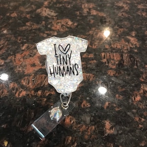 May include: A silver glitter badge holder with a white baby onesie design that says "I love tiny humans".