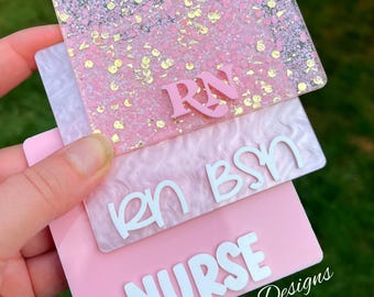 May include: A stack of pink and white nurse badge holders. The top badge is clear with pink glitter and gold flecks, with the letters "RN" in pink. The second badge is white with the letters "RN BSN". The bottom badge is pink with the word "NURSE" and the text "Bushel & Peck Designs".