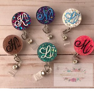 May include: A selection of glittery badge reels with various coloured backgrounds, each displaying a unique initial in a cursive font. The reels are round and have a retractable cord and a clip on the back.