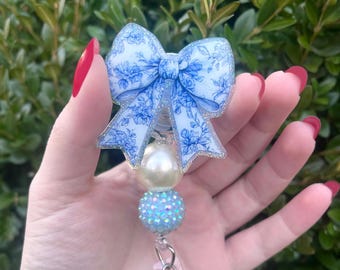 May include: A blue and white floral patterned bow with glitter accents, attached to a white pearl and a blue iridescent rhinestone bead. The bow is attached to a clear plastic retractable badge holder.