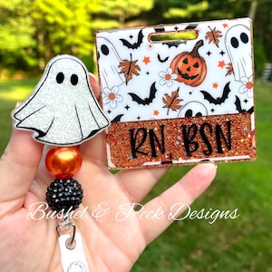 May include: Halloween-themed badge reel with a white glitter ghost charm and a badge holder featuring a pumpkin, bats, and ghosts. The badge holder has the text "RN BSN" on an orange glitter background. The reel has orange and black beads.